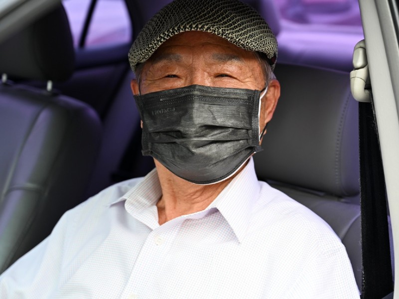 O coreano Il Han Bae está sentado dentro do carro, no banco do motorista. Ele está máscara preta e boina xadrez